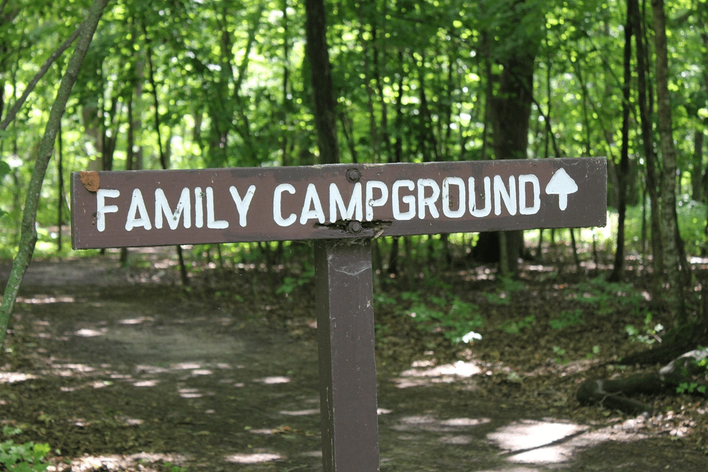 Wooden signpost with white painted letters reading Family Campground and an arrow pointing forward placed along a dirt path surrounded by green forest trees