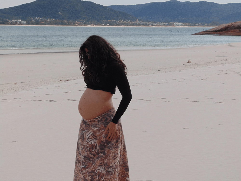 Pregnant woman standing barefoot on a quiet sandy beach gently holding her skirt and looking down at her baby bump with ocean waves and mountains in the background